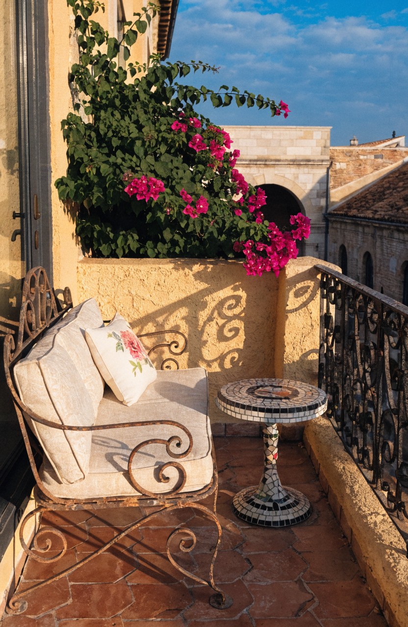 Wrought iron armchair with white cushions and small round mosaic table on terracotta-tiled balcony surrounded by lush green plants and cascading pink bougainvillea over black metal railing, yellow stucco wall, and view of beige stone arches under blue sky.