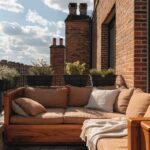 A beige-cushioned wooden sofa with a light blanket on a deck balcony next to a glass door, with potted succulents and plants against a brick wall and two chimneys under a blue sky.