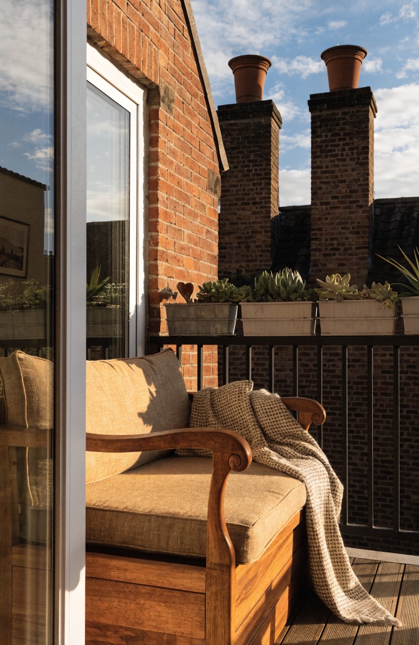 A beige-cushioned wooden sofa with a light blanket on a deck balcony next to a glass door, with potted succulents and plants against a brick wall and two chimneys under a blue sky.