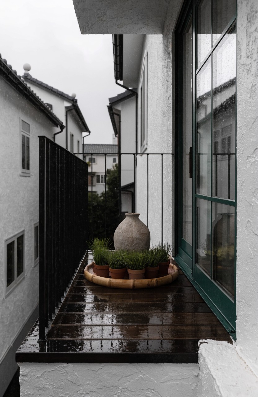 Wet wooden balcony floor with black railing, large gray stone pot containing green plants inside a bamboo ring, next to green sliding glass doors on a white stucco house exterior on a rainy day.
