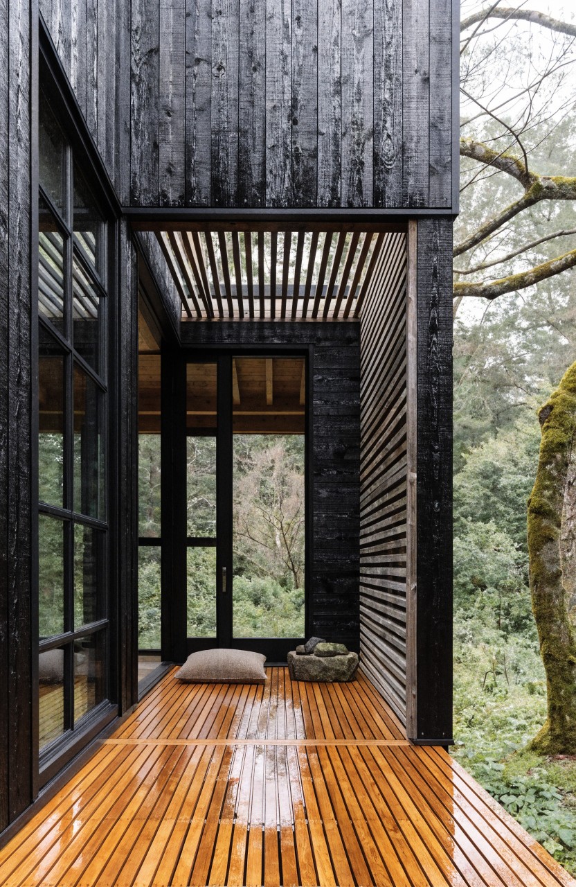 Black wood-clad modern house exterior with a warm-toned wooden deck balcony, large black-framed glass doors, slatted wood screen, floor cushion, basket, and surrounding trees.