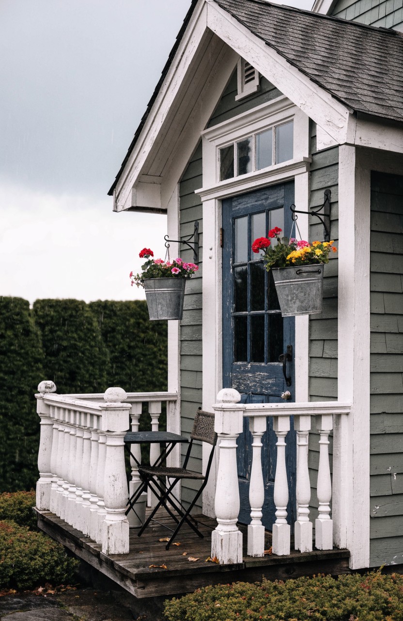 Small green clapboard gabled structure with white trim, dark blue door, white porch railing holding a table and chair, red geraniums in galvanized bucket planters hanging from posts, hedges around the base.