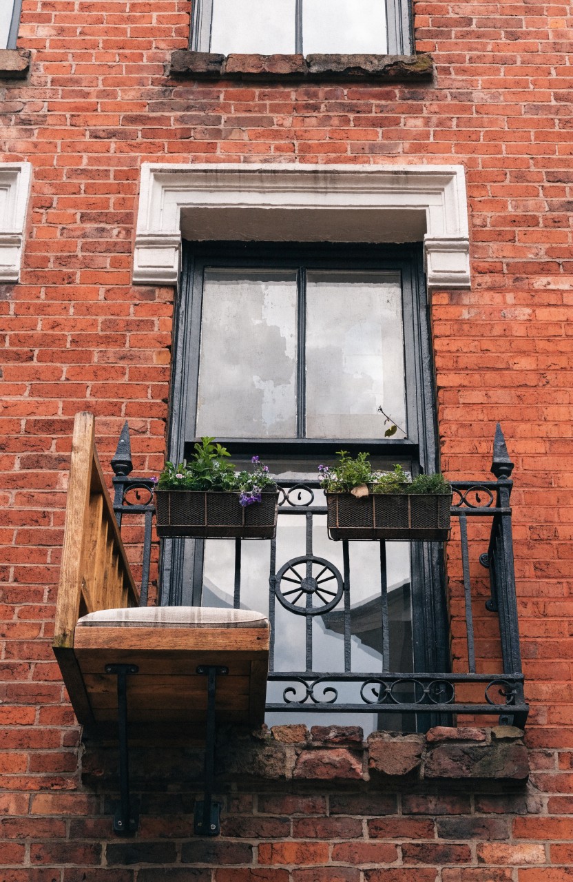 Narrow black wrought-iron balcony on red brick wall with wooden chair, flower boxes containing purple flowers and greenery, and large dark-framed window above.