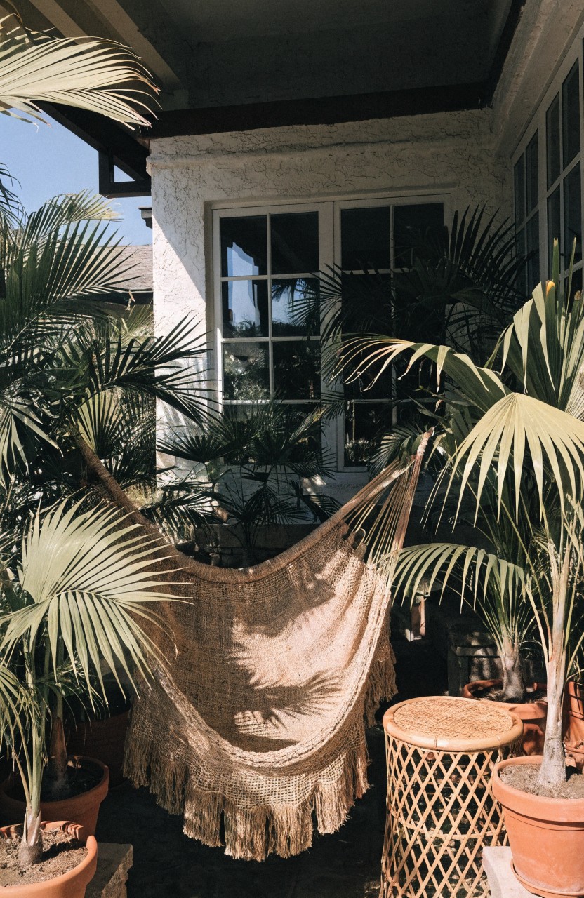 A beige fringed hammock hangs from the ceiling of a covered balcony surrounded by large potted palm plants, with a rattan stool nearby, white stucco walls, and large multipane windows.