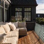 Modern black house with wooden balcony deck, beige L-shaped outdoor sofa with cushions, tall potted grasses, and glass railings with metal accents.