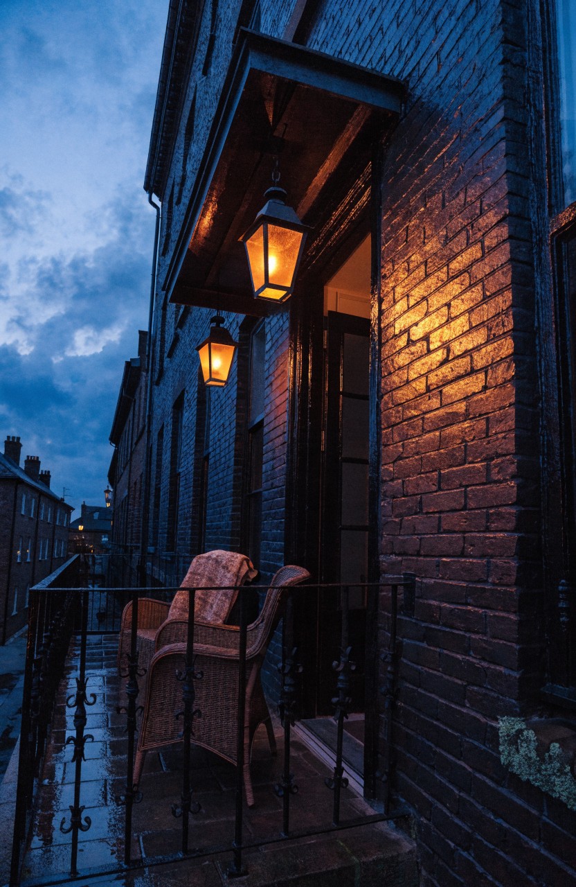 Dark brick townhouse exterior at dusk with a second-floor balcony railing holding two wicker armchairs, one draped in a plaid blanket, two hanging lanterns lit warmly, and an open door below showing interior light.