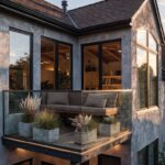 A contemporary stone-clad house exterior with a curved balcony featuring glass railings, wooden decking, potted grasses and plants, and LED strip lights glowing underneath the railings at dusk.