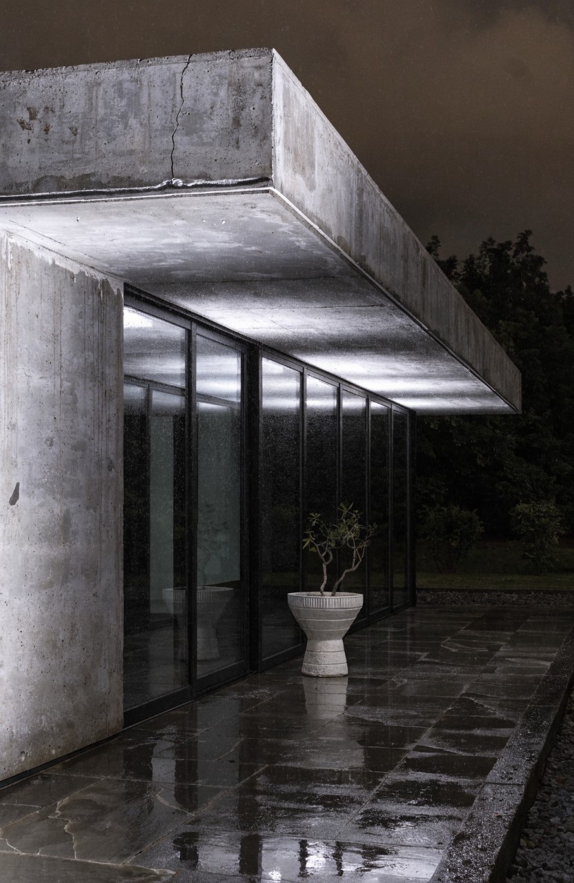 Night view of concrete house exterior with cantilevered roof edge lit by linear white LED strips, shining on glass doors, wet stone patio, and potted plant in white pedestal pot.