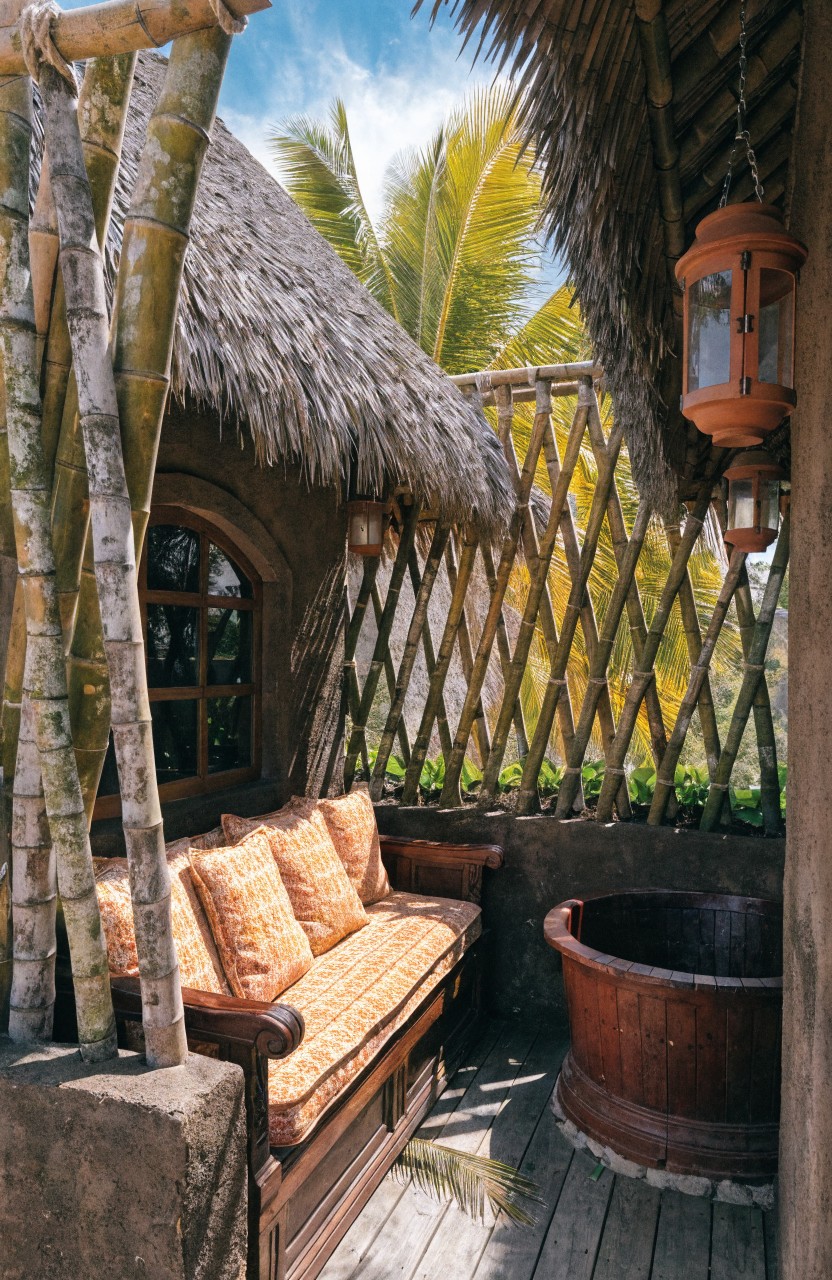 Balcony lounge under a thatched roof with bamboo supports and lattice screening, orange bench cushions, wooden tub, hanging lanterns, and palm trees nearby.
