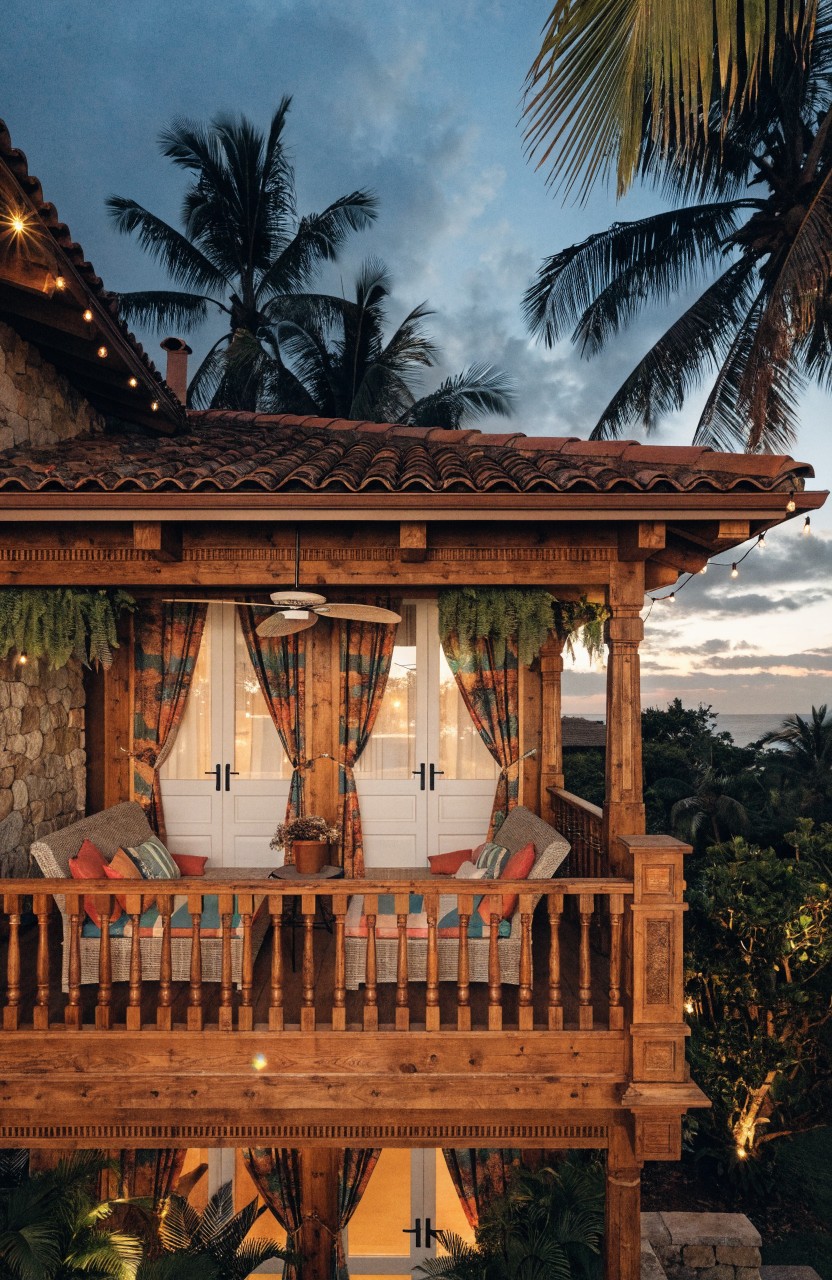 Elevated wooden balcony on a stone tropical house with colorful cushions on benches, double white doors, hanging plants, palm trees surrounding it at dusk with string lights.