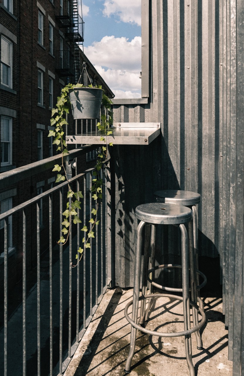 Small urban balcony with two silver metal bar stools at a narrow protruding metal shelf, ivy trailing from a hanging bucket and railing, corrugated gray metal wall, black railing, and views of brick buildings under partly cloudy sky.
