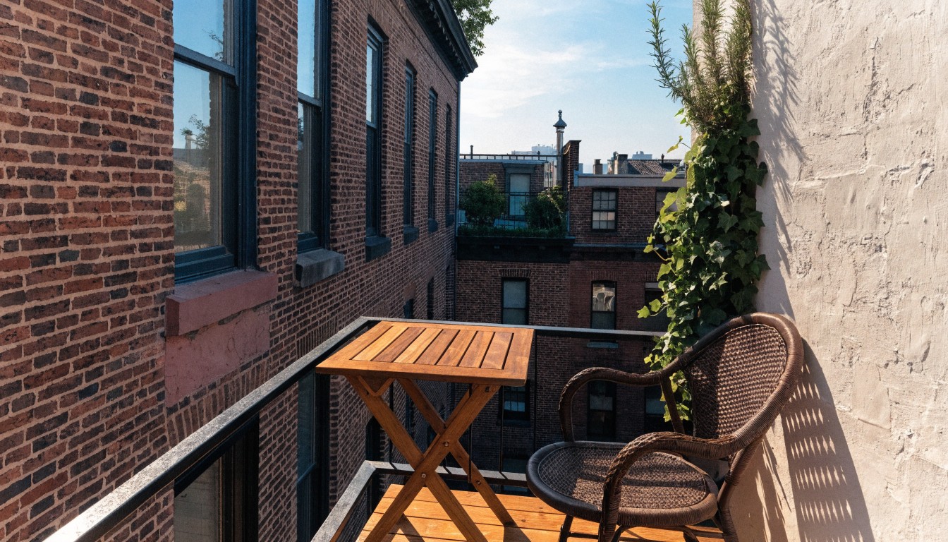 Small corner balcony on a red brick building with wooden decking, black metal railing, wicker armchair, folding wooden table, potted rosemary, and ivy overflowing from a white planter box.