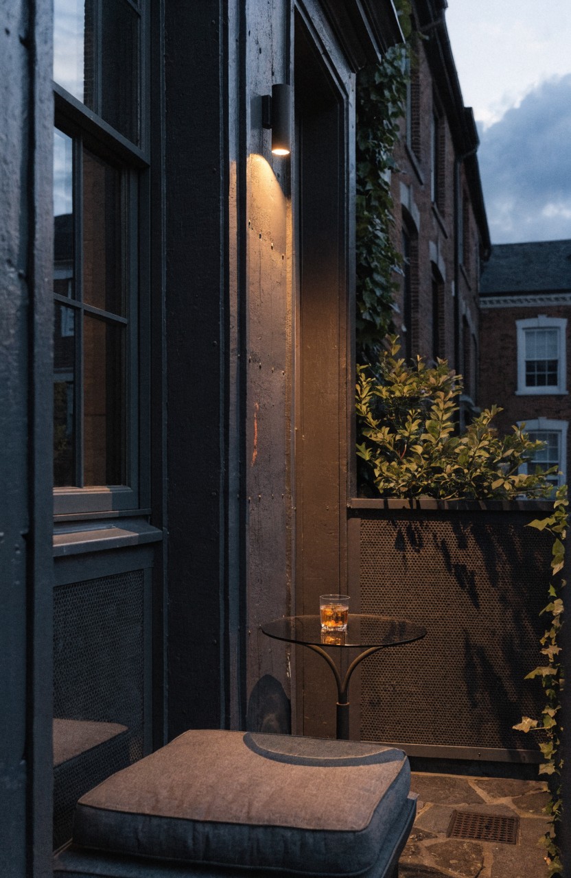 Narrow balcony at dusk with two wall-mounted sconces lighting a cushioned floor seat, small glass table holding a drink, ivy on railings and walls, and brick building exterior.