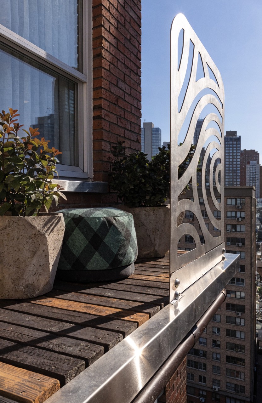 Small urban balcony featuring wooden deck flooring, two stone planters with greenery, a green pouf stool, and a tall wavy perforated metal screen railing against a brick building with cityscape beyond.