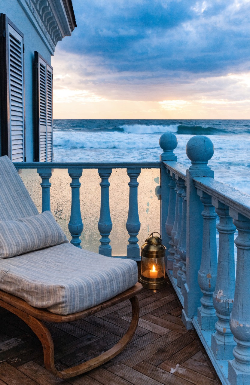 Wooden chaise lounge chair with light beige striped cushions and cushions on a balcony deck with blue painted railings overlooking ocean waves at dusk with a lit brass lantern nearby.