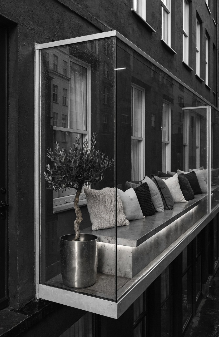 Black and white photo of a rectangular glass-enclosed balcony protruding from a dark brick multi-story building, with a cushioned bench seat, potted olive tree, and underlit ledge.