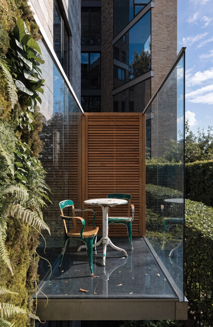 Narrow balcony with dense vertical green plant walls on both sides, wooden slatted privacy screen, glass railings, dark flooring, small round table, and two colorful chairs.