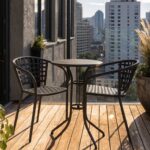 A small outdoor balcony deck with two large weathered concrete pots of tall feathery grasses and leafy plants flanking a round black metal table and two matching latticed chairs with beige cushions, overlooking a city skyline.