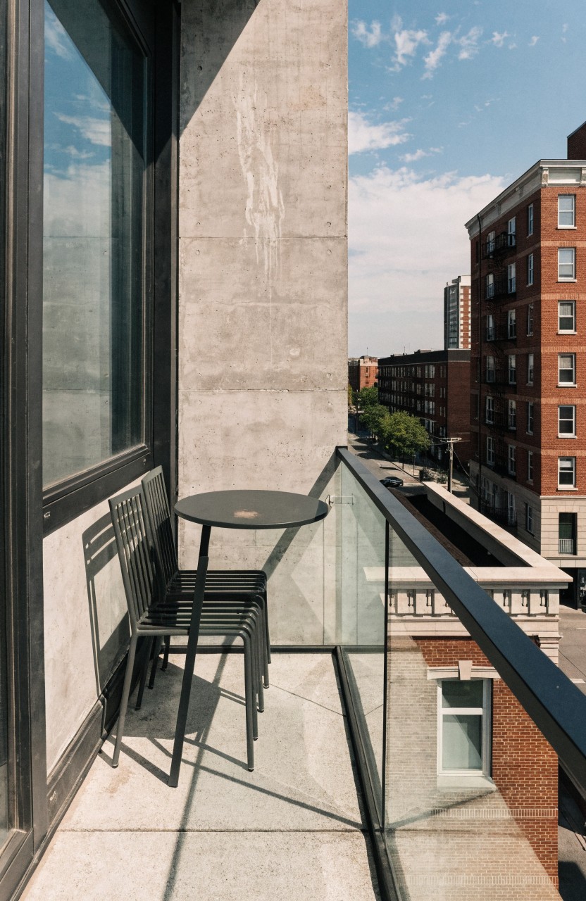 Narrow concrete balcony with a small round black table and two black metal chairs against the wall, glass railing, overlooking brick buildings, trees, and city street.