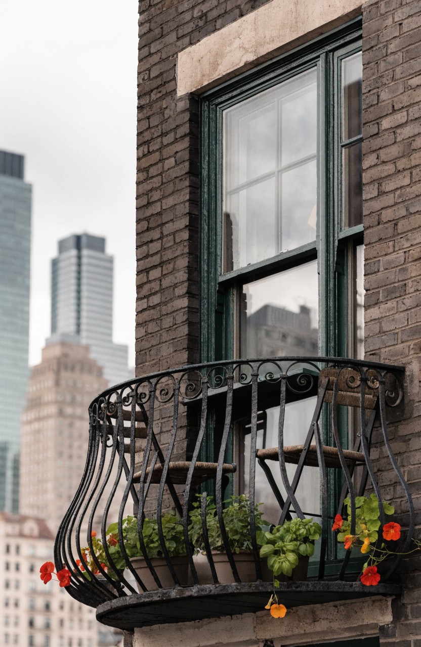 Narrow black wrought-iron balcony on a brick building exterior with two small wooden chairs around a tiny table, potted plants including red flowers and herbs, and a green window above, city skyline visible behind.