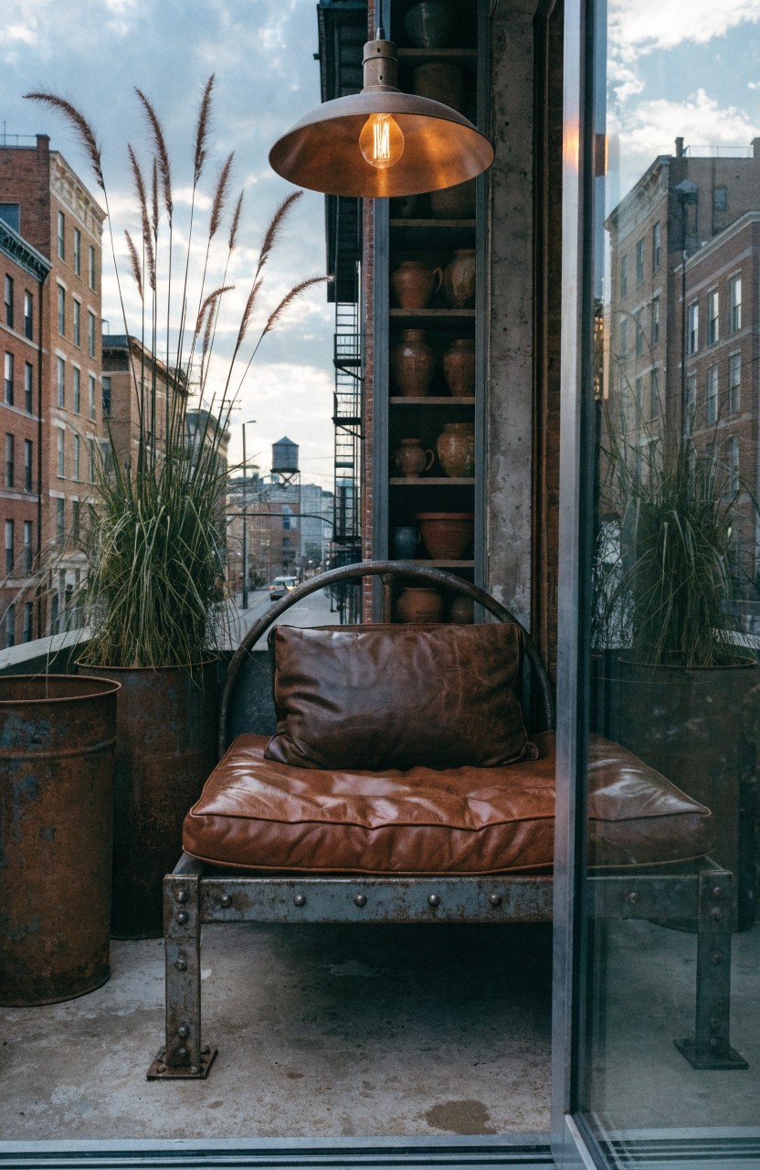 Apartment balcony viewed through glass doors with a low metal-framed daybed piled with brown leather cushions, flanked by tall pampas grass in large pots and rusty metal drums, terracotta pots on wooden shelves, and a hanging pendant light over urban brick buildings.