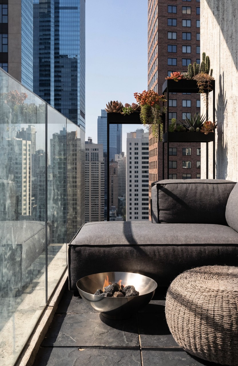 Apartment balcony with glass railing, dark tiled floor, gray L-shaped sofa, woven round pouf, shallow metal fire bowl containing small flames, black shelves holding potted succulents and plants, brick wall backdrop, and city skyscrapers visible.