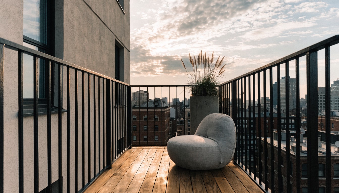 Balcony with wooden deck floor, black metal railing, white pod-shaped cushioned chair, tall potted pampas grass next to it, glass door, and city buildings in the background under partly cloudy sky.