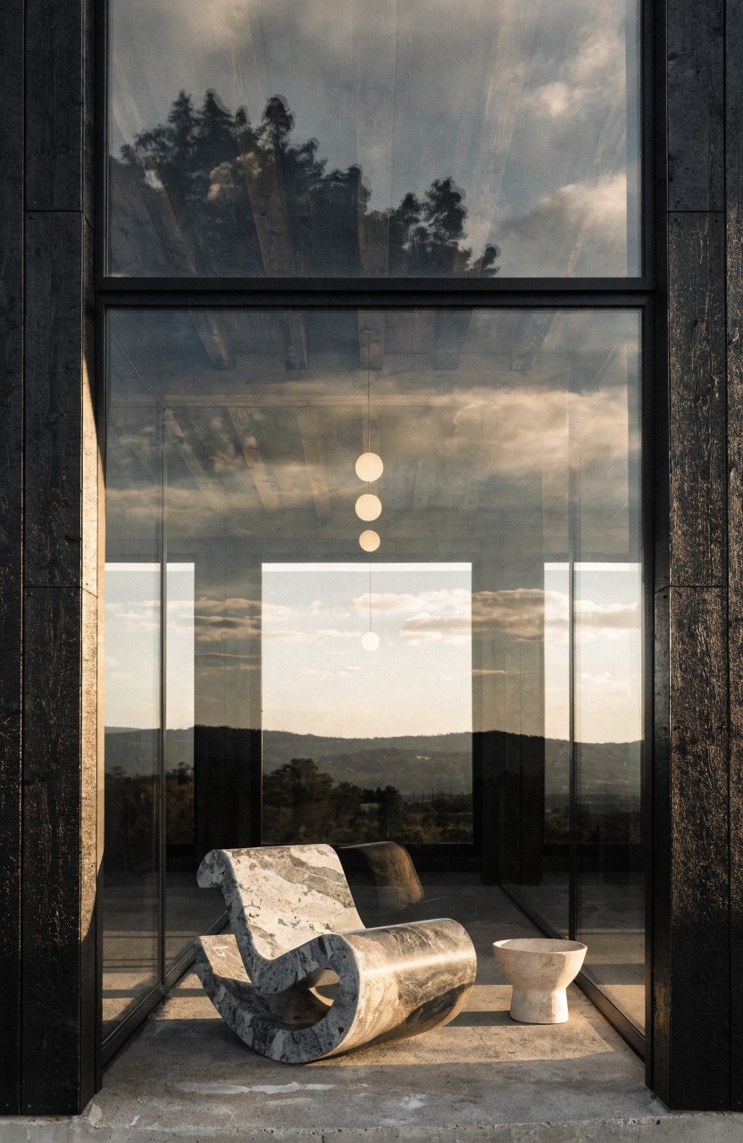 Curved gray lounge chair and white cylindrical stool on a concrete terrace in front of tall black metal-framed glass panels overlooking trees and hills at sunset.