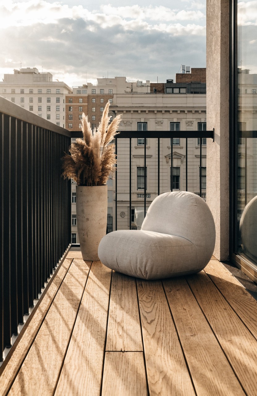 Balcony with wooden deck floor, black metal railing, white pod-shaped cushioned chair, tall potted pampas grass next to it, glass door, and city buildings in the background under partly cloudy sky.