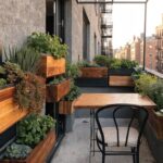 Narrow balcony with wooden planter boxes mounted vertically along the exterior walls at varying heights, filled with herbs, greens, and succulents, plus a small wooden table and chair under a glass canopy.
