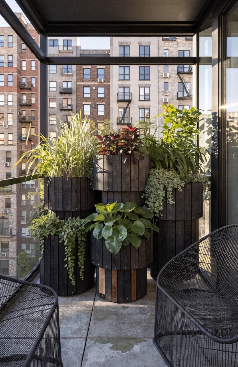 A compact balcony enclosed by glass railing holds several tall black wooden planters at varying heights filled with green leafy plants, flanked by black wire chairs, overlooking tall city buildings.