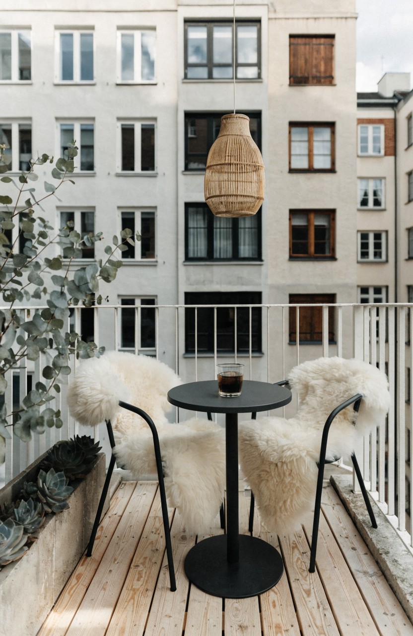 Wooden balcony deck with white metal railing holding two white sheepskin-covered armchairs around a small black round table topped with a coffee cup, potted plants on the ledge, rattan hanging lamp, and city apartments in the background.