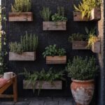 A narrow urban balcony with a dark wall covered in wooden wall-mounted planters filled with greenery, a tall climbing vine with white flowers, a cushioned wooden bench, and a large terracotta pot next to brick buildings under a partly cloudy sky.