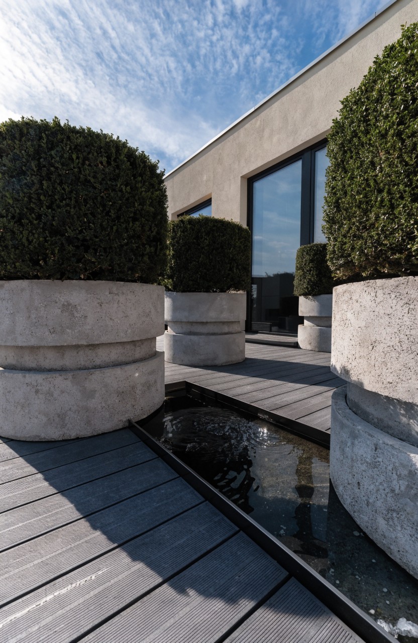 Wooden deck balcony with four large cylindrical beige concrete planters holding trimmed spherical boxwood shrubs, a narrow water channel along the deck edge, and a beige building facade featuring dark-framed glass doors.