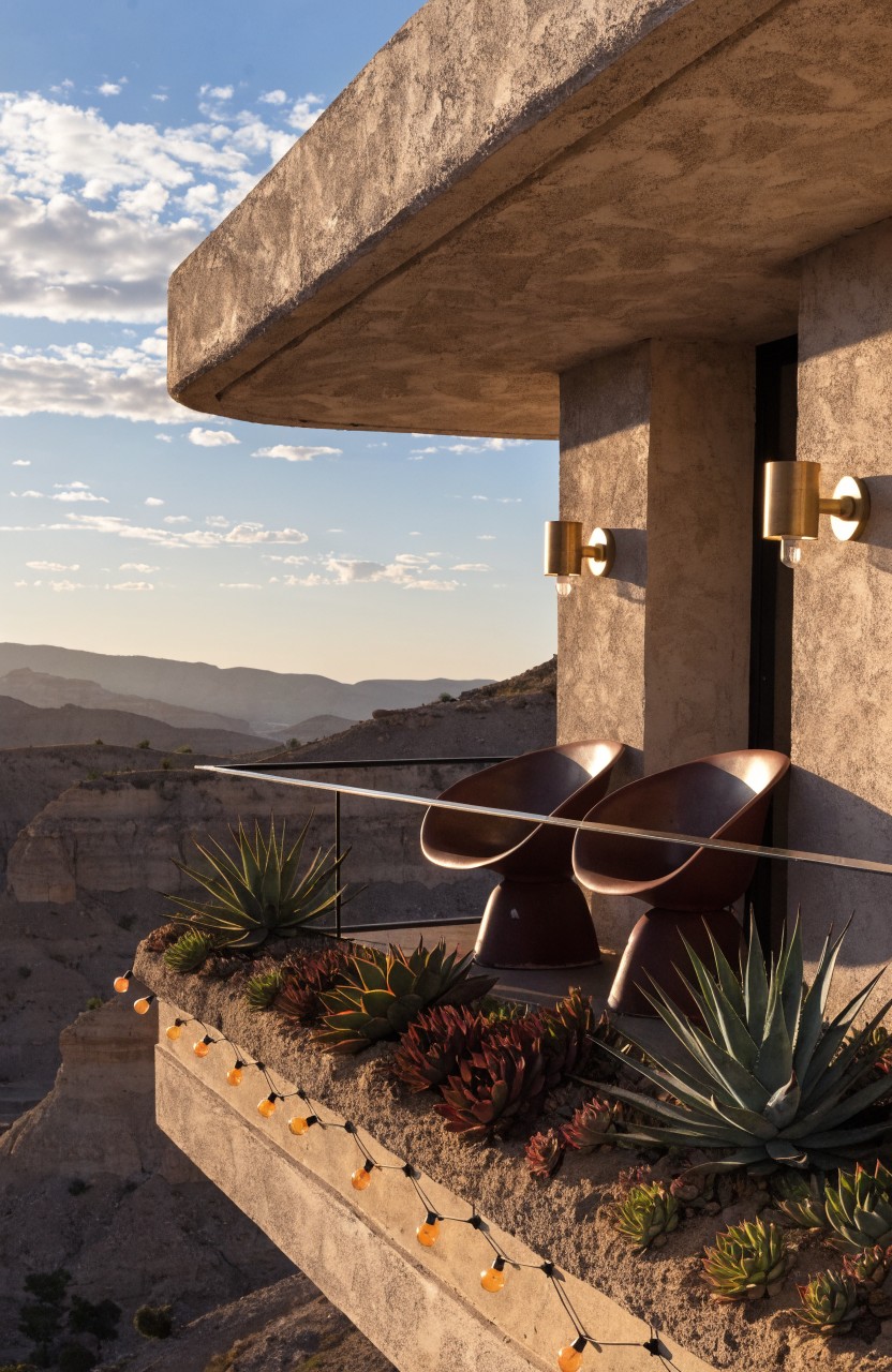 Cantilevered concrete balcony edge with succulent planters, string lights draped along them, two brown chairs, gold wall sconces beside doors, and desert canyon view at sunset.