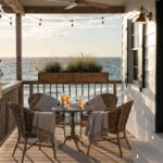 White clapboard house exterior with a wooden deck balcony over rocky oceanfront, featuring a pergola strung with pendant lights, potted lavender plants along the railing, a small round table, two wicker chairs draped with a blanket, and sliding glass doors.