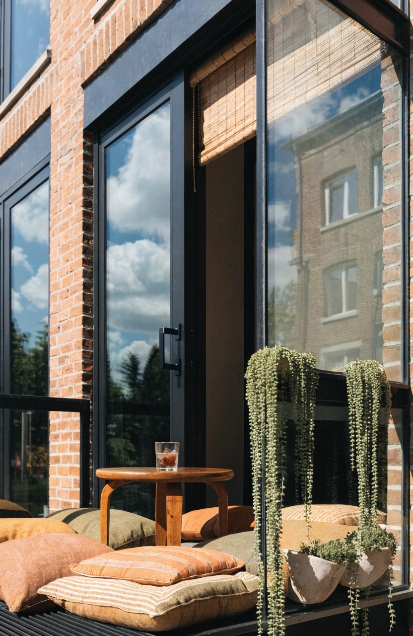 Balcony with orange and beige floor cushions piled around a low wooden table holding a glass of iced drink, white pots of trailing green plants hanging from the edge, and black sliding glass doors with bamboo shades against brick walls.