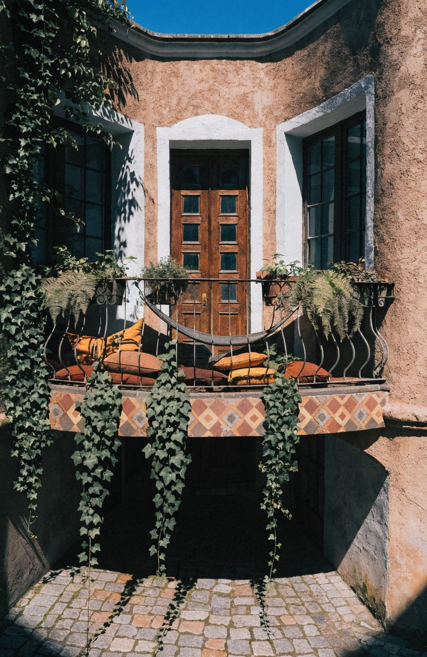 Corner of a beige stucco house with ivy climbing the walls, white-trimmed windows, a wooden double door, wrought-iron balcony railing holding potted plants and a hammock swing with orange cushions, terracotta tiled floor, and cobblestone path below.