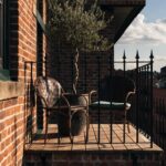 Brick townhouse exterior with black wrought iron balcony railing holding two wicker chairs, a small round table, and a potted olive tree beside large multipane windows.