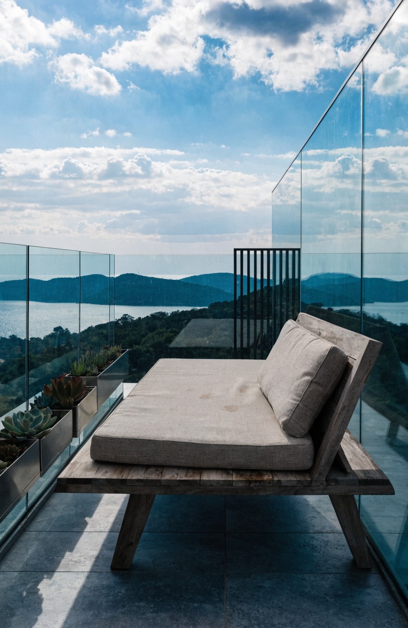 A low wooden bench with beige cushions sits on a modern balcony with glass railings, metal planters holding succulents, and a view of the sea and distant mountains under partly cloudy skies.
