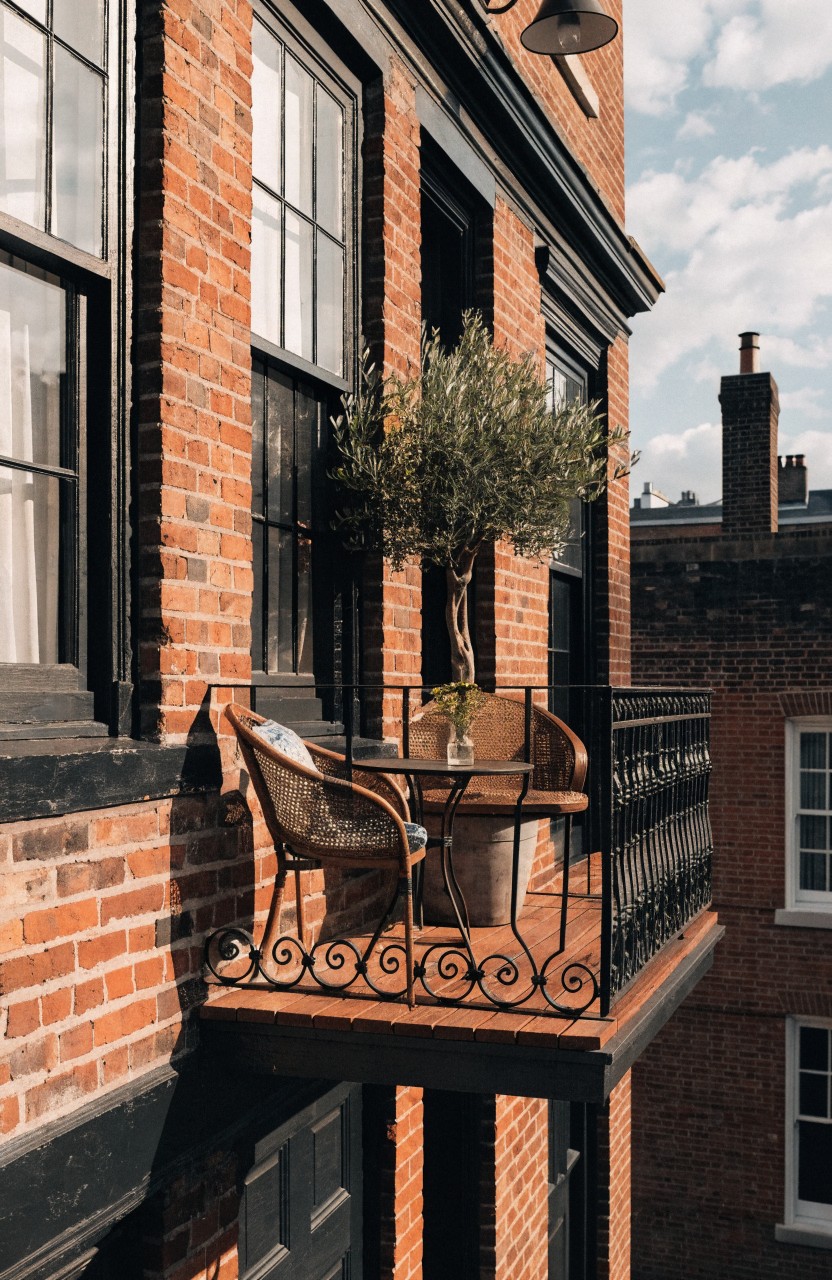 Brick townhouse exterior with black wrought iron balcony railing holding two wicker chairs, a small round table, and a potted olive tree beside large multipane windows.