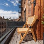 Wooden cantilevered lounge chair mounted on brick wall of a balcony with glass railing, wooden decking, slatted wood screen, black planter with plant, and globe wall light.