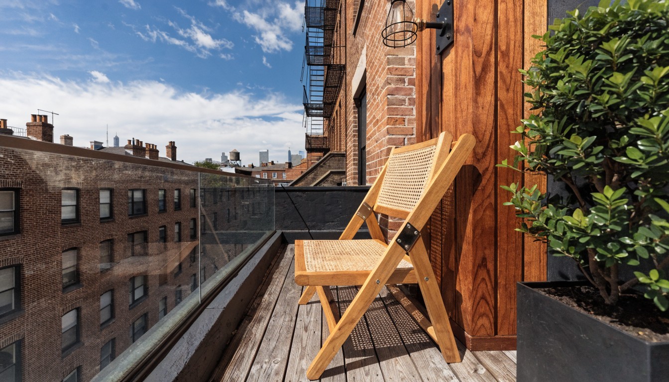 Wooden cantilevered lounge chair mounted on brick wall of a balcony with glass railing, wooden decking, slatted wood screen, black planter with plant, and globe wall light.
