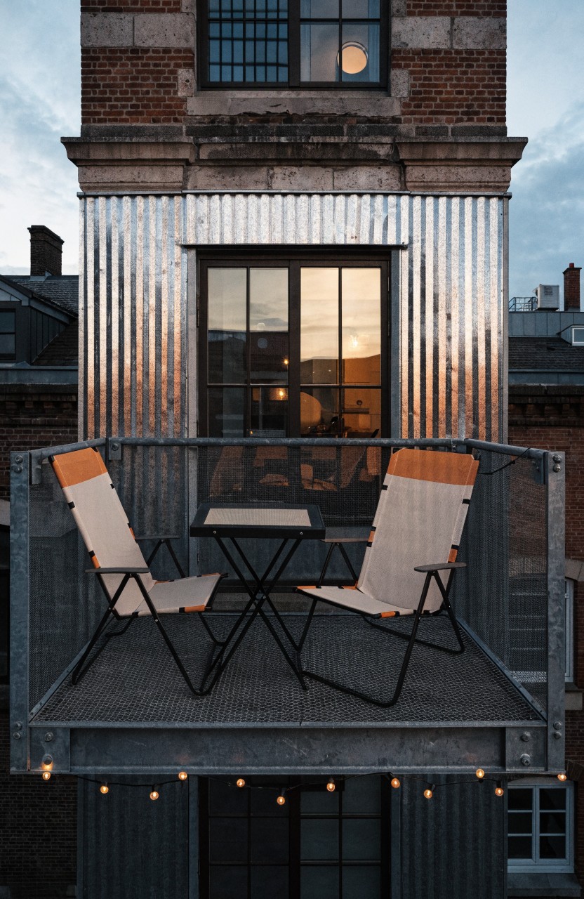 Narrow exterior balcony on a brick building with corrugated metal siding, two orange and white folding chairs, a small table, large windows, and string lights below.