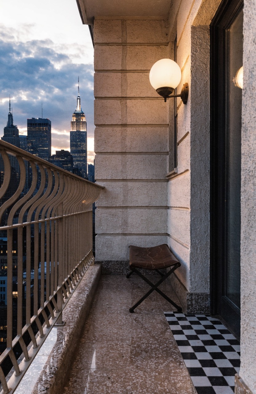 Narrow exterior balcony with beige stone walls, black wrought iron railing, wall-mounted white globe light, small tan leather stool, and city skyline view at dusk.