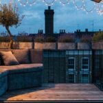 Black metal balcony railing with wooden decking, built-in gray fabric bench with cushions, potted plants like olive tree and lavender, string fairy lights overhead, and glass doors to indoor space at dusk with city buildings in background.