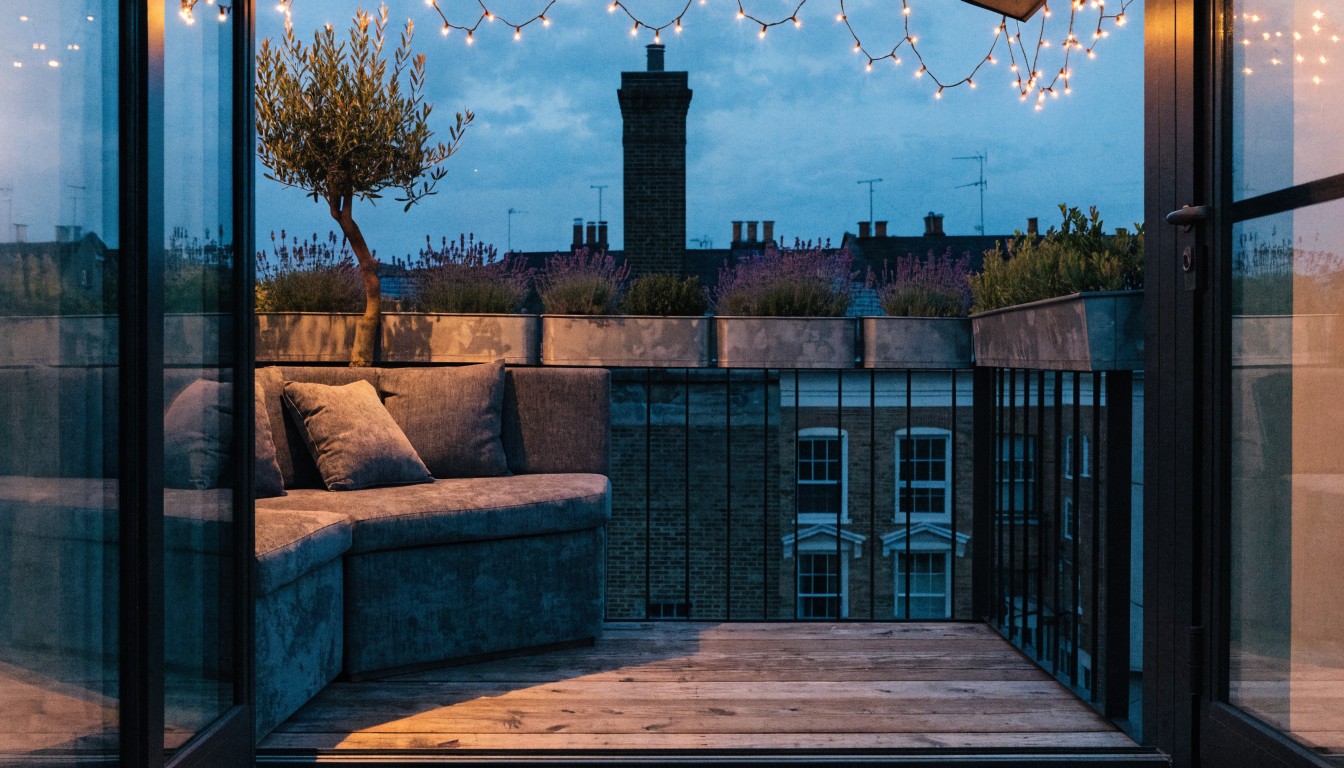 Black metal balcony railing with wooden decking, built-in gray fabric bench with cushions, potted plants like olive tree and lavender, string fairy lights overhead, and glass doors to indoor space at dusk with city buildings in background.
