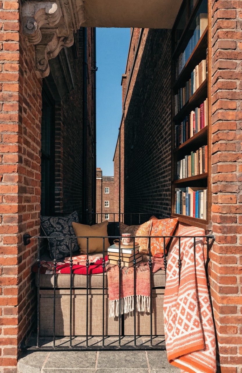 Narrow brick balcony passageway with cushioned bench piled with colorful pillows, blankets, and books, flanked by bookshelves and a metal railing.