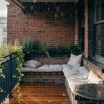 Corner balcony on brick building in the rain with wooden deck floor, wrought iron railing, cushioned wraparound bench around round metal table, and potted rosemary and ivy plants.