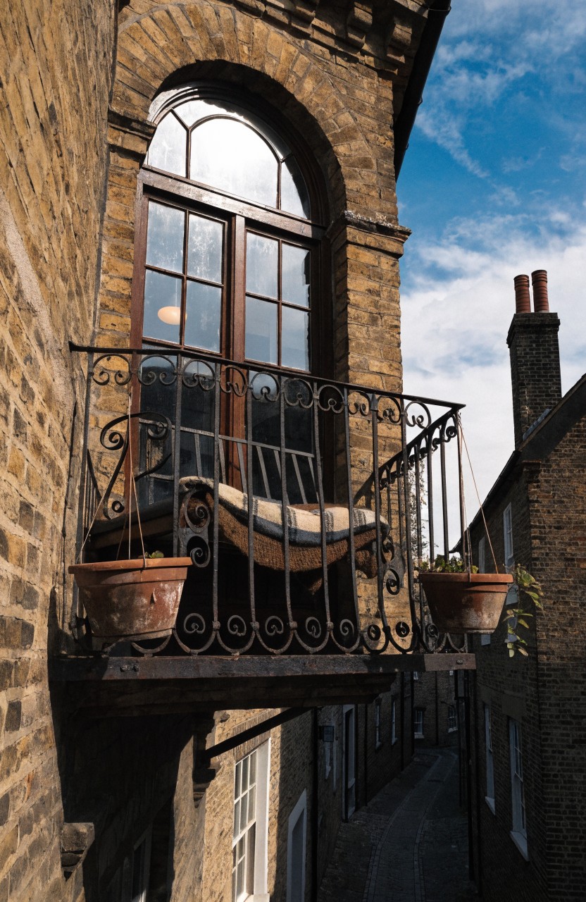 Wrought iron balcony on a beige brick townhouse with arched window holds a cushioned white hanging seat and two terracotta planters with green plants.